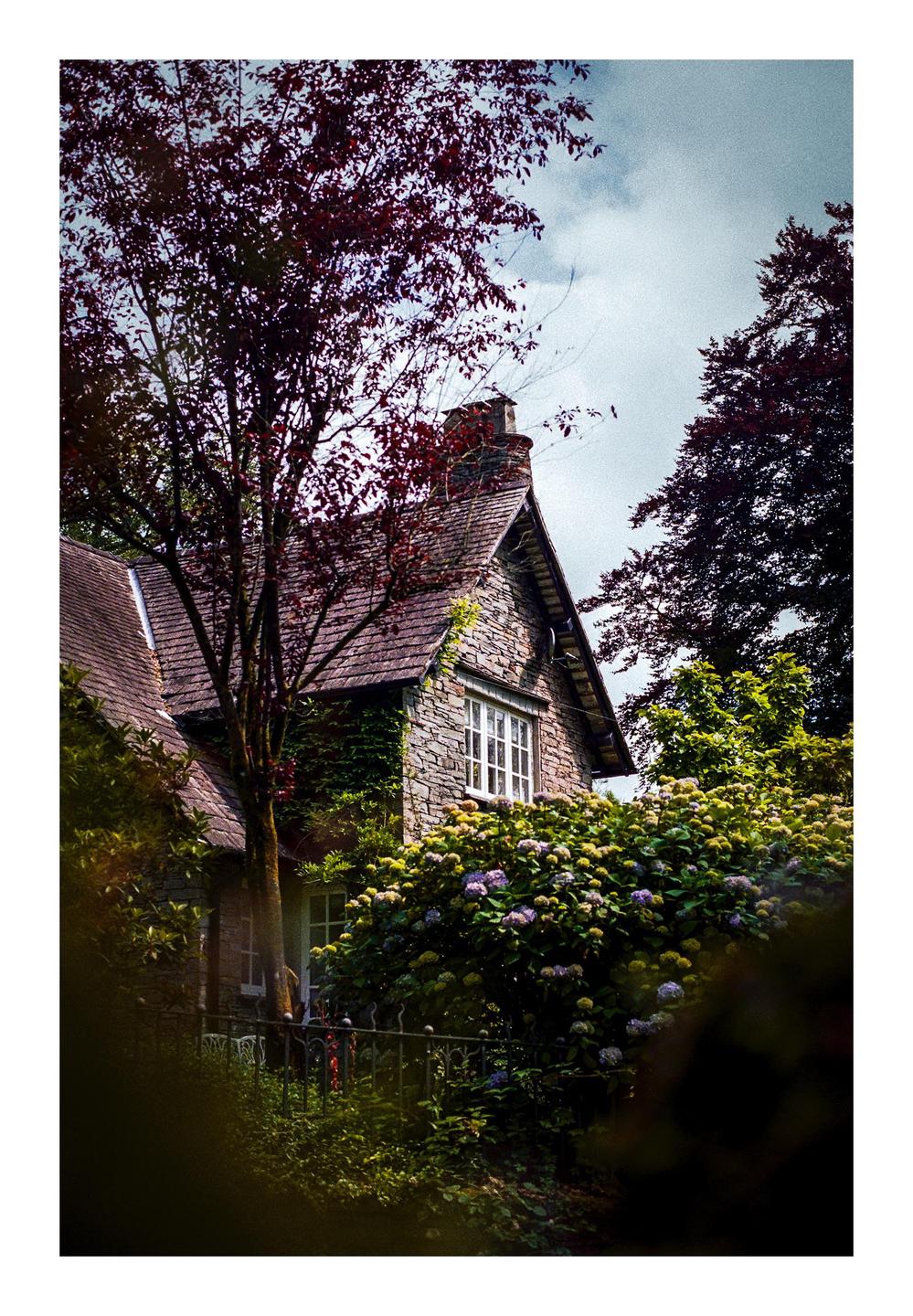 A stone cottage with a white window is surrounded by trees and flowering bushes under a cloudy sky.