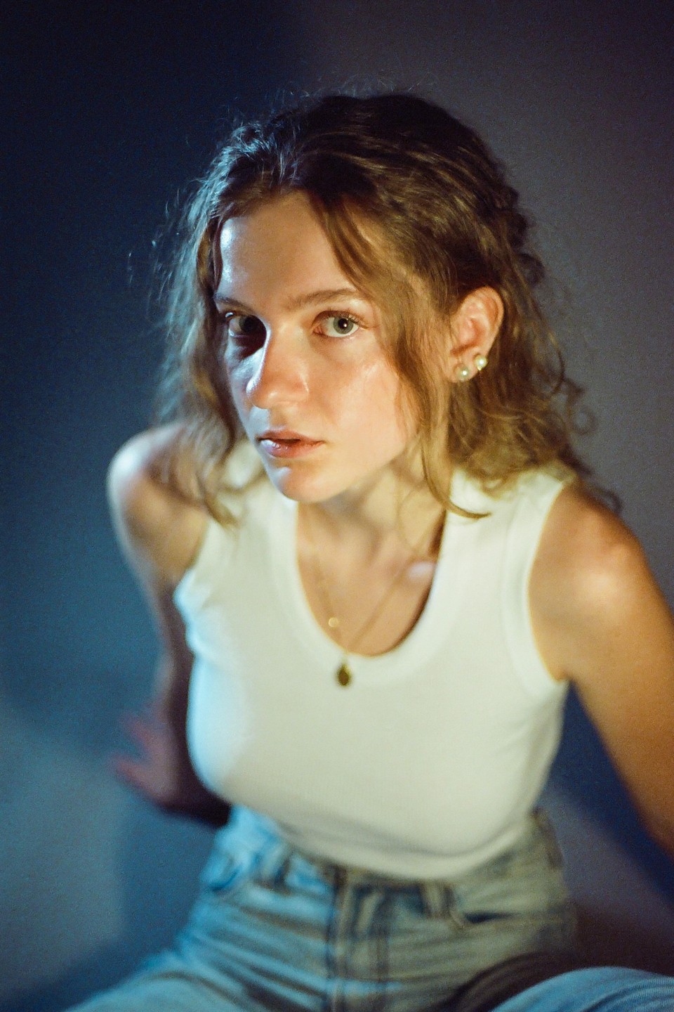 A woman with curly hair and a white tank top looks directly at the viewer.