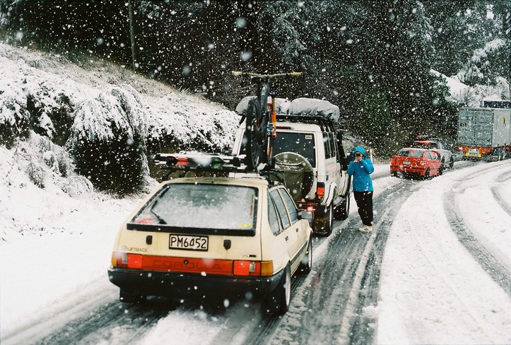 A person in a blue jacket stands on a snowy road next to a white car with a bicycle on top, while snow falls.