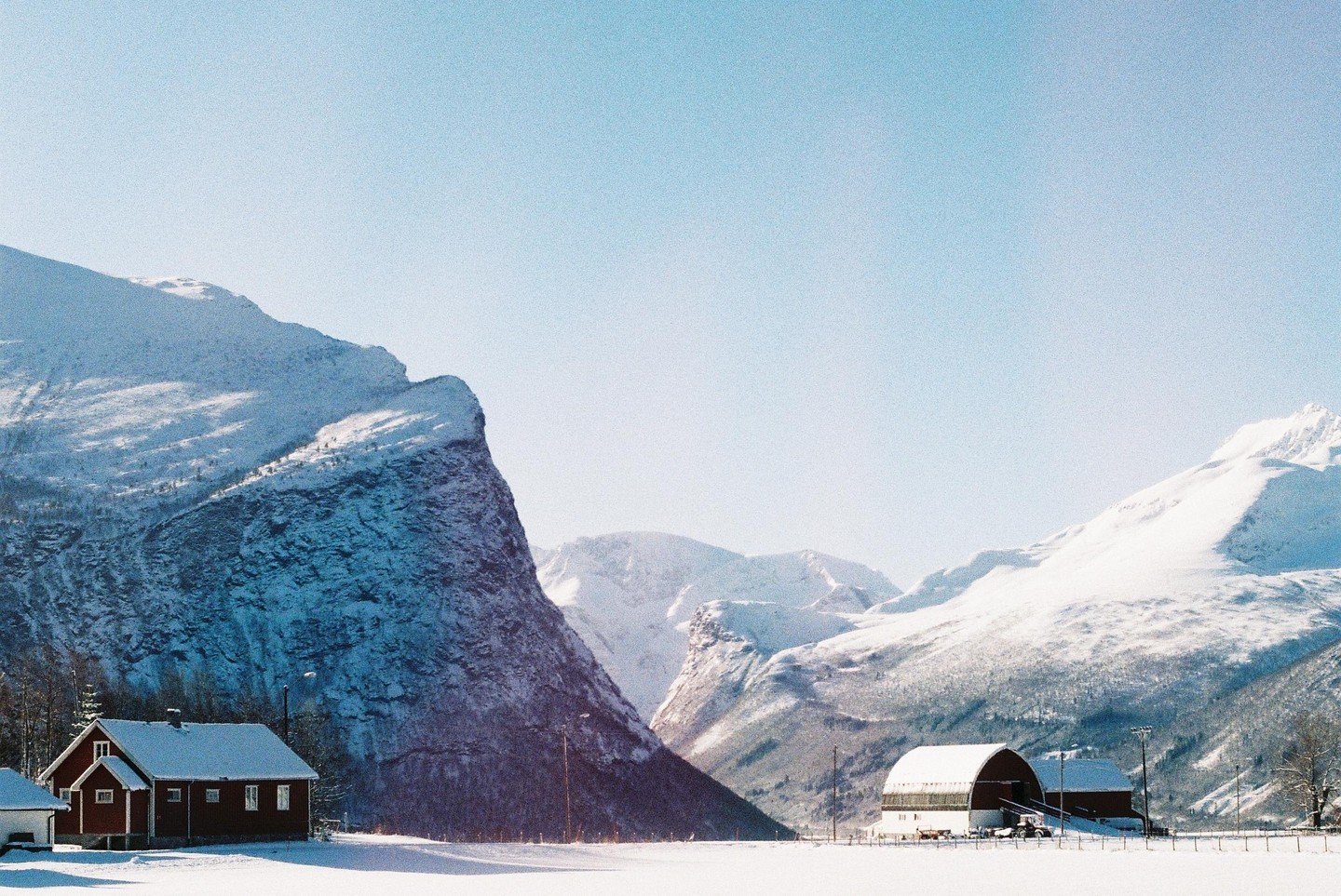 A red house and a barn sit in a snow-covered valley surrounded by large, snow-capped mountains under a clear sky.
