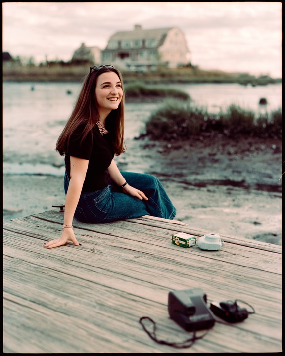 A young woman sits on a wooden dock next to a body of water with a large house in the background.
