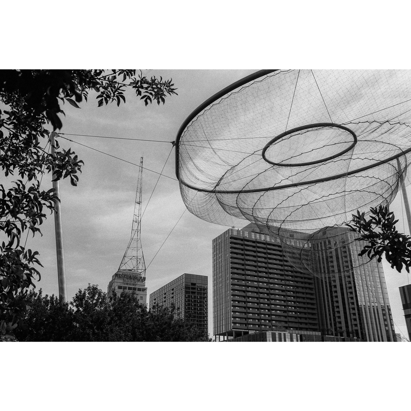 A large, circular, netted sculpture hangs over buildings and trees in a city.