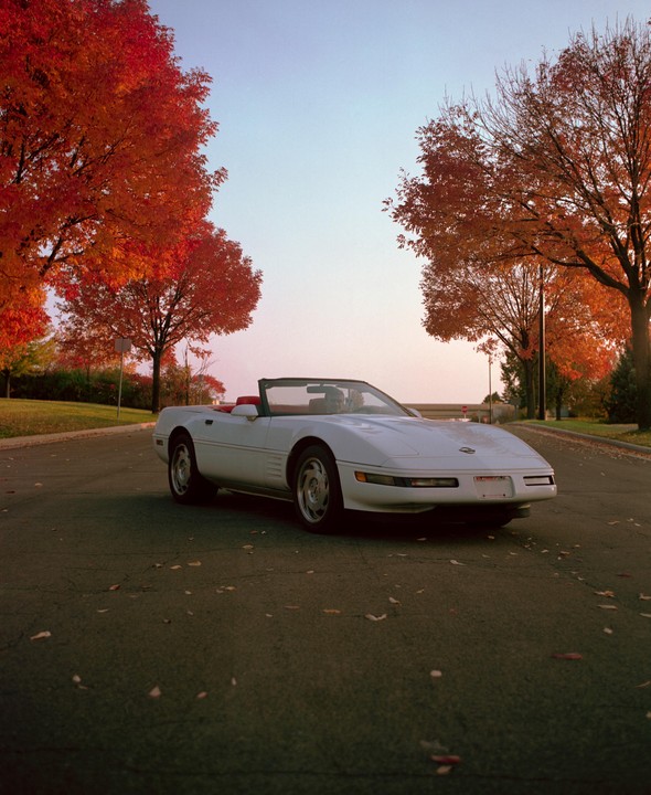 A white convertible car is parked on an asphalt road lined with trees displaying autumn foliage.