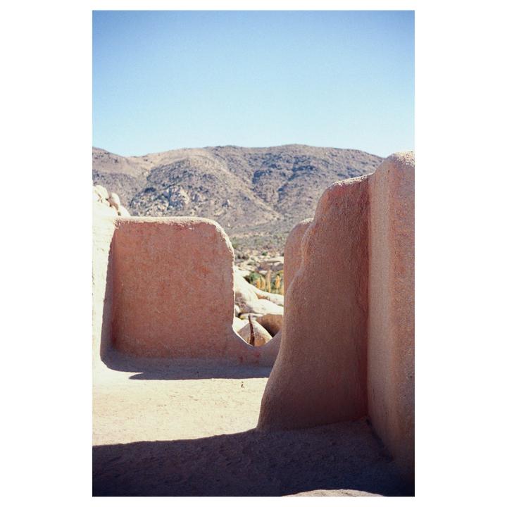 A desert landscape with mountains in the background is framed by two adobe walls in the foreground.