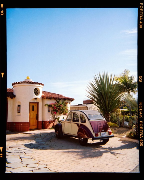A two-tone car is parked on a paved driveway in front of a white house with a red tile roof and a palm tree.