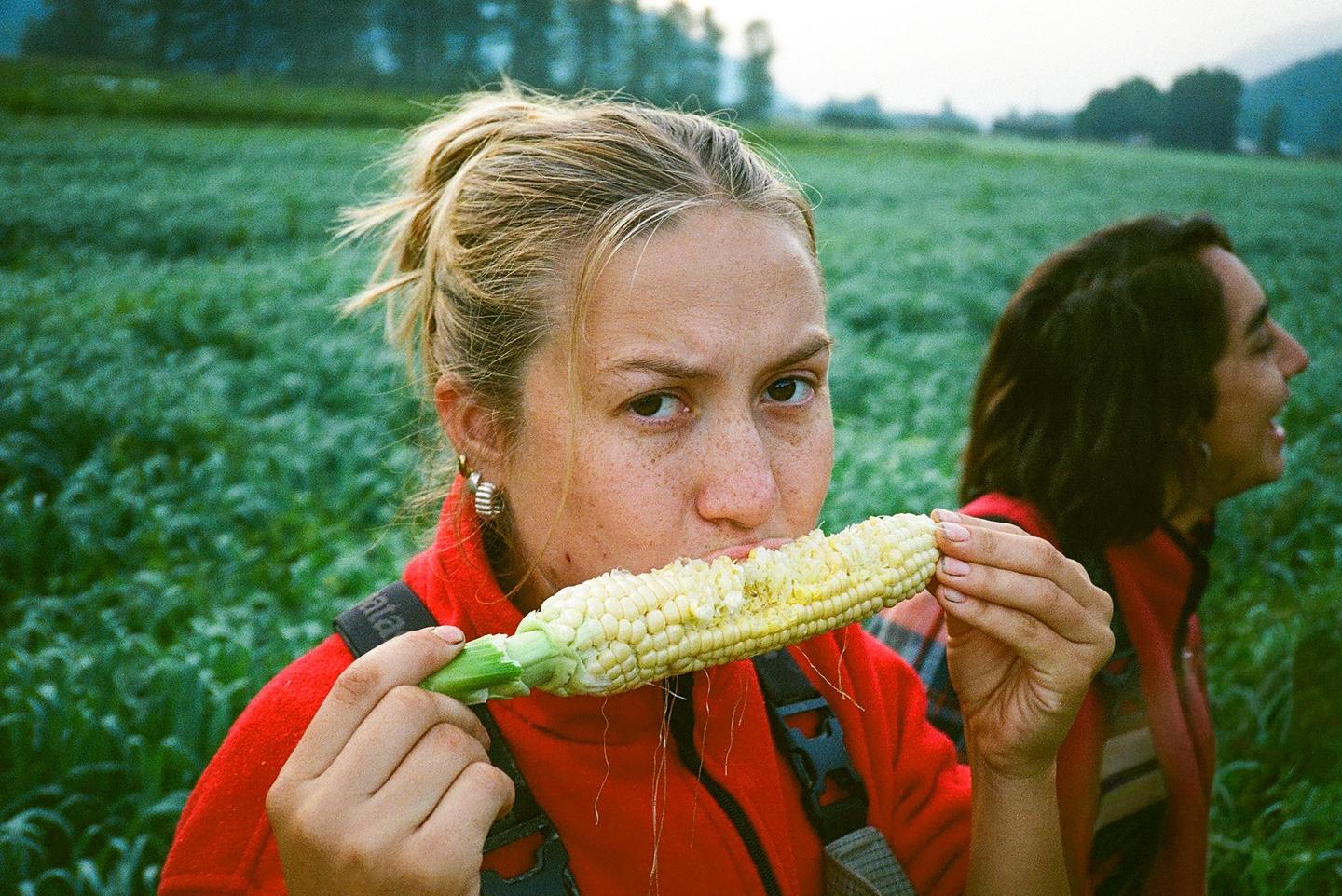A woman in a red jacket eats an ear of corn in a field with another person in the background.