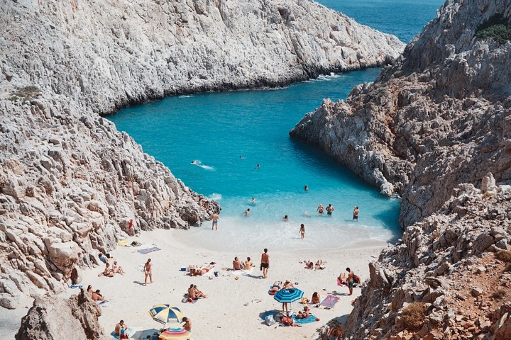 A crowd of people relax on a white sand beach surrounded by rocky cliffs and turquoise water.