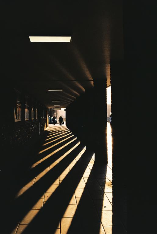 Two people walk through a dark underpass with strong shadows cast across the ground.