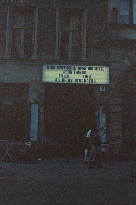 A movie theater marquee advertises several films above a dark entrance where two people stand.