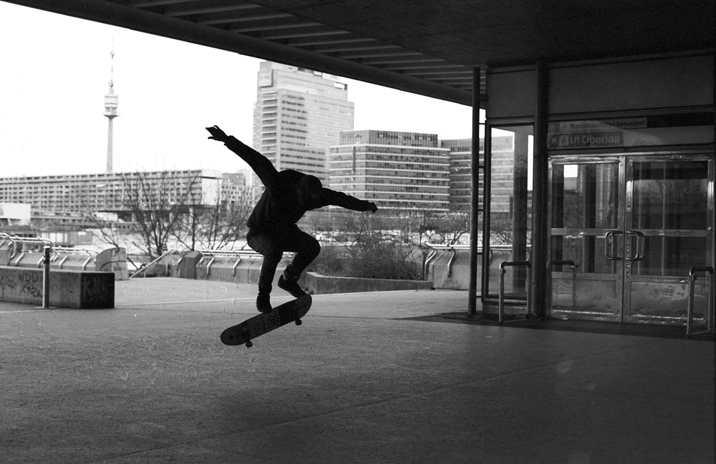 A skateboarder performs a trick in an urban setting with buildings and a tower in the background.