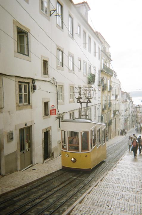 A yellow tram travels up a steep, narrow street lined with white buildings and people walking on the sidewalk.