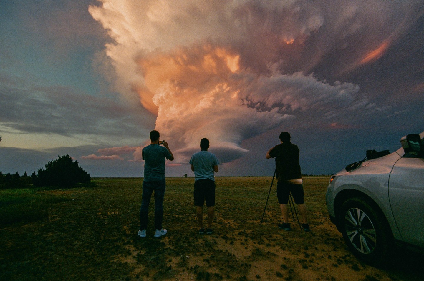 Three men stand in a field looking at a large, dramatic cloud formation in the sky.