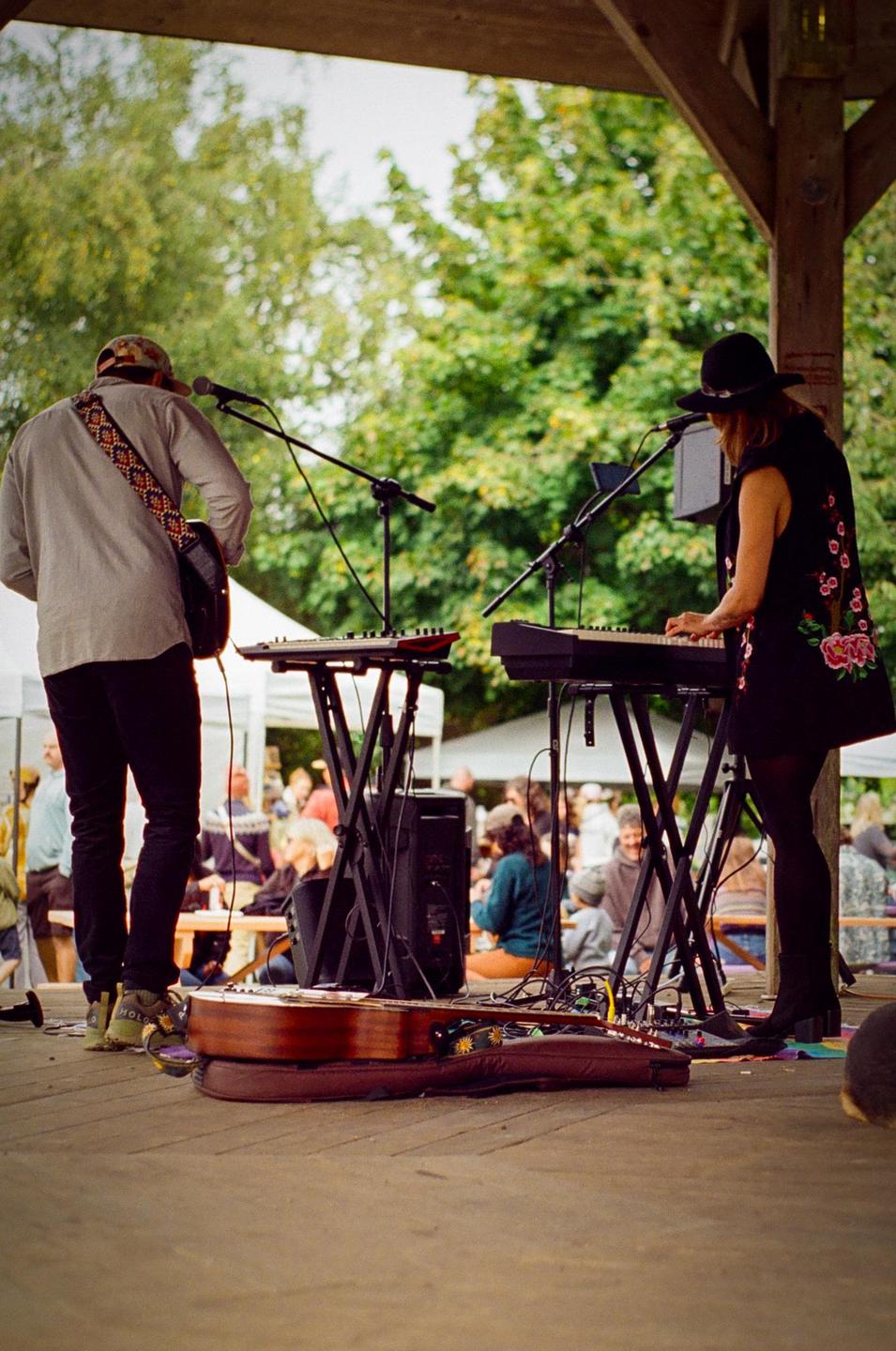 A man and a woman perform music on a stage outdoors with an audience in the background.