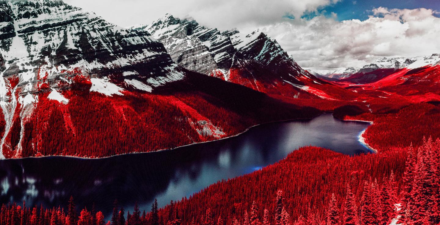A lake is surrounded by red trees and snow-capped mountains under a cloudy sky.
