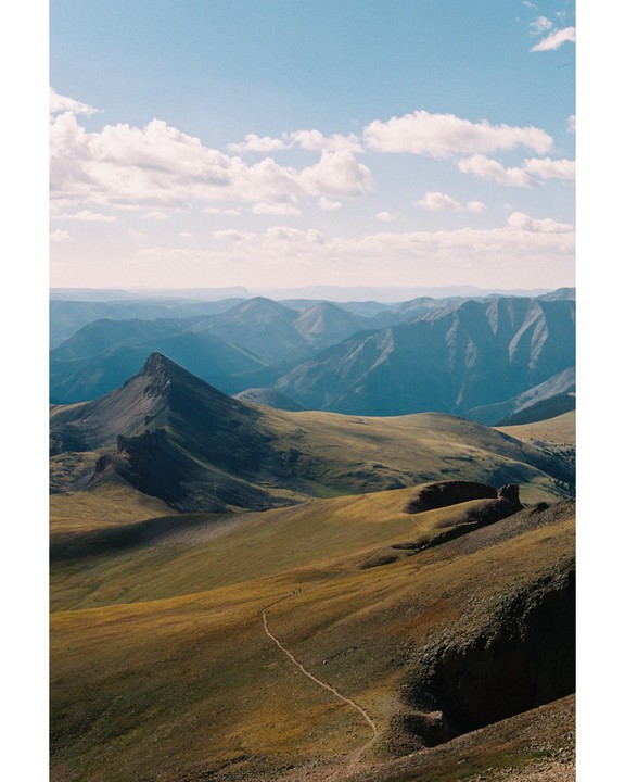 A narrow trail winds through a vast mountain landscape under a partly cloudy sky.
