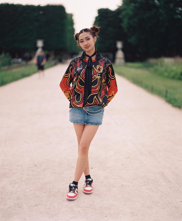 A woman wearing a flame-patterned jacket and denim skirt stands on a gravel path.