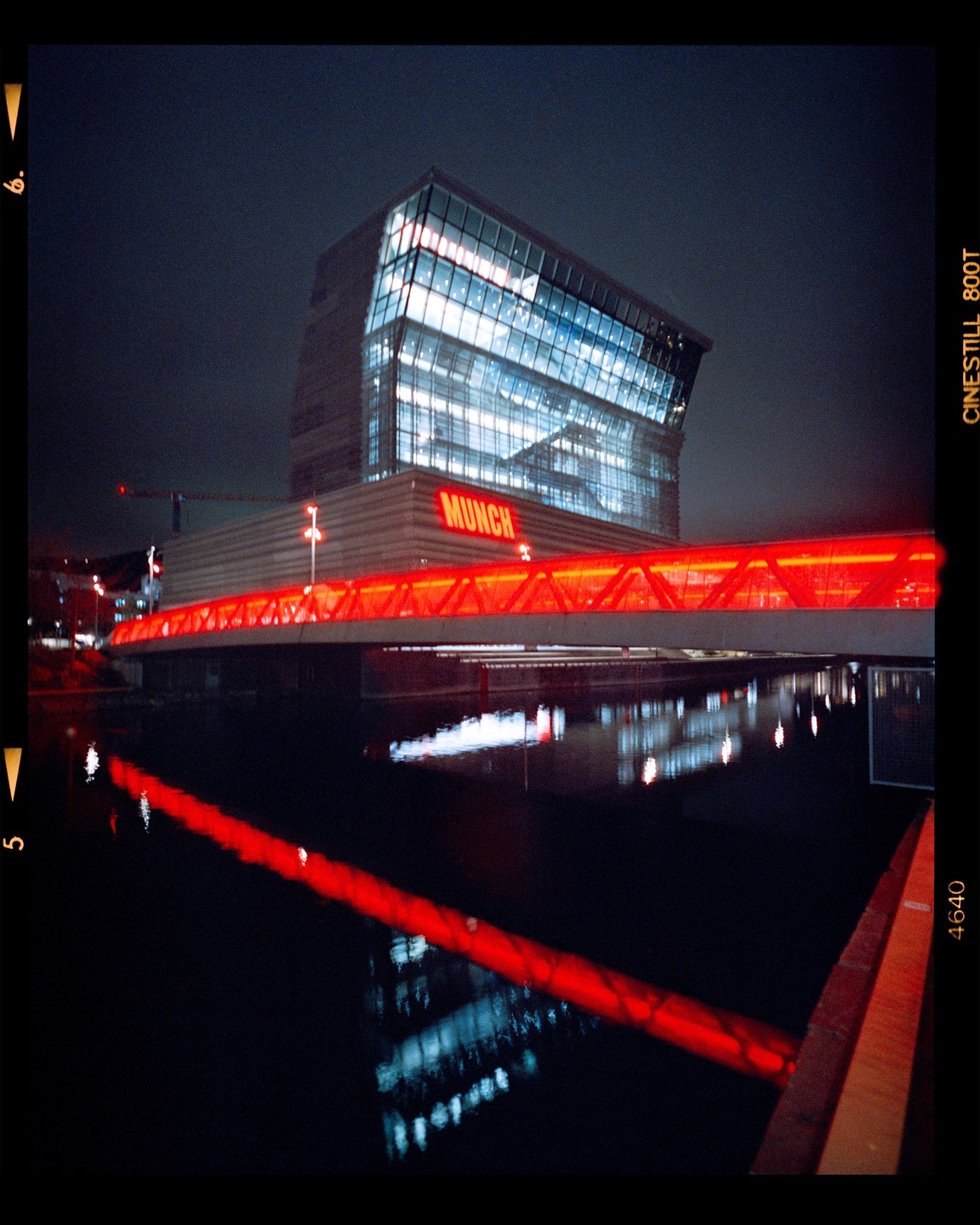 A modern building with a red bridge and its reflection in the water at night.