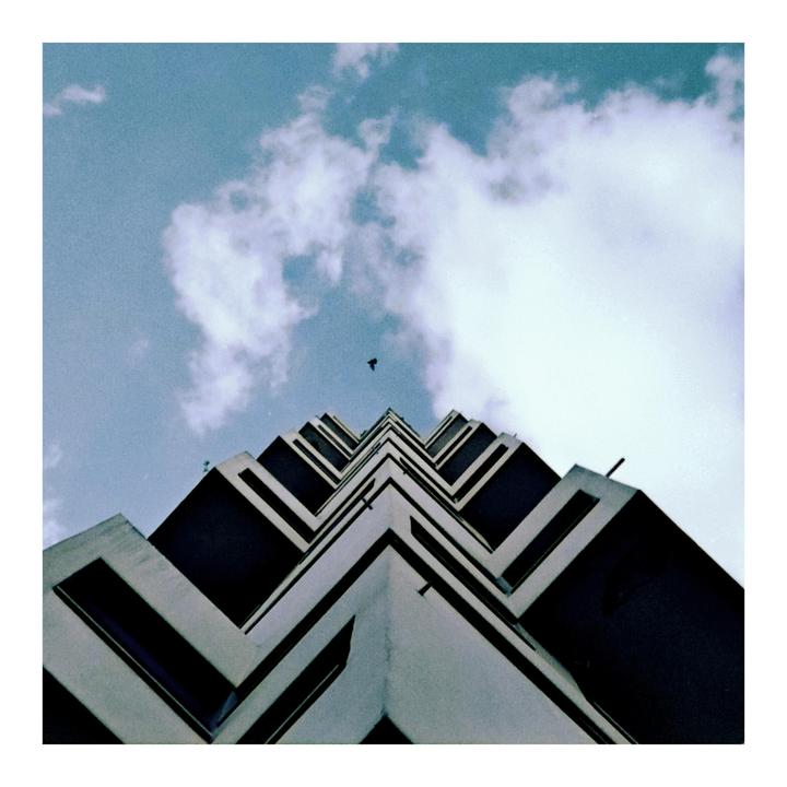 A low-angle view of a building with a zig-zag pattern on its facade, with a bird flying in the cloudy sky above.