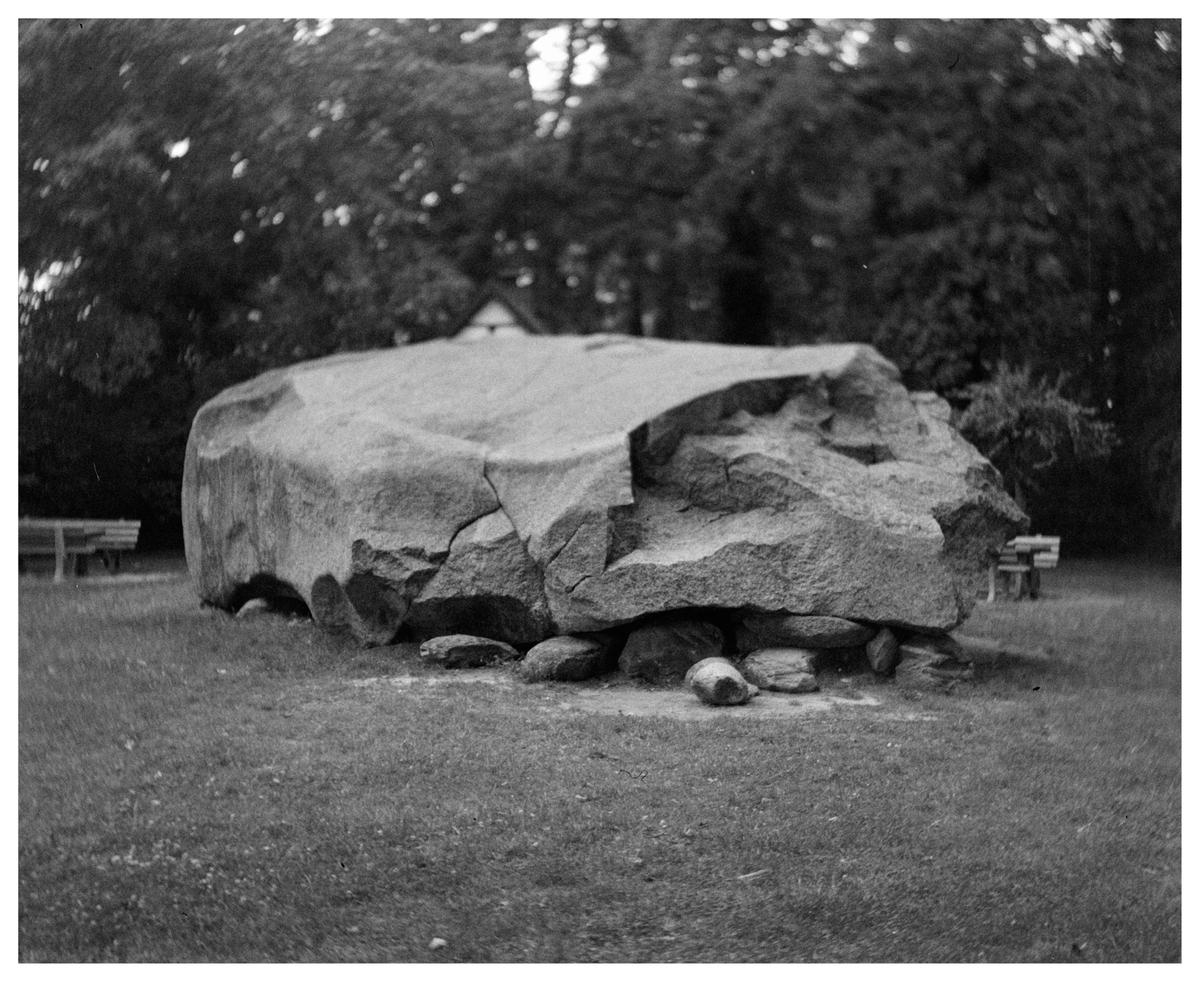A large, cracked boulder rests on smaller stones in a grassy area with trees and benches in the background.