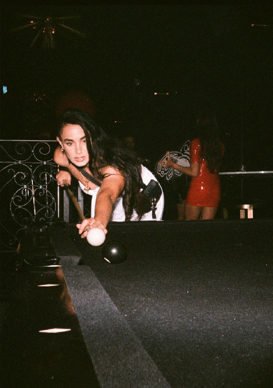 A woman with long dark hair leans over a pool table, holding a cue stick and aiming at a white cue ball.