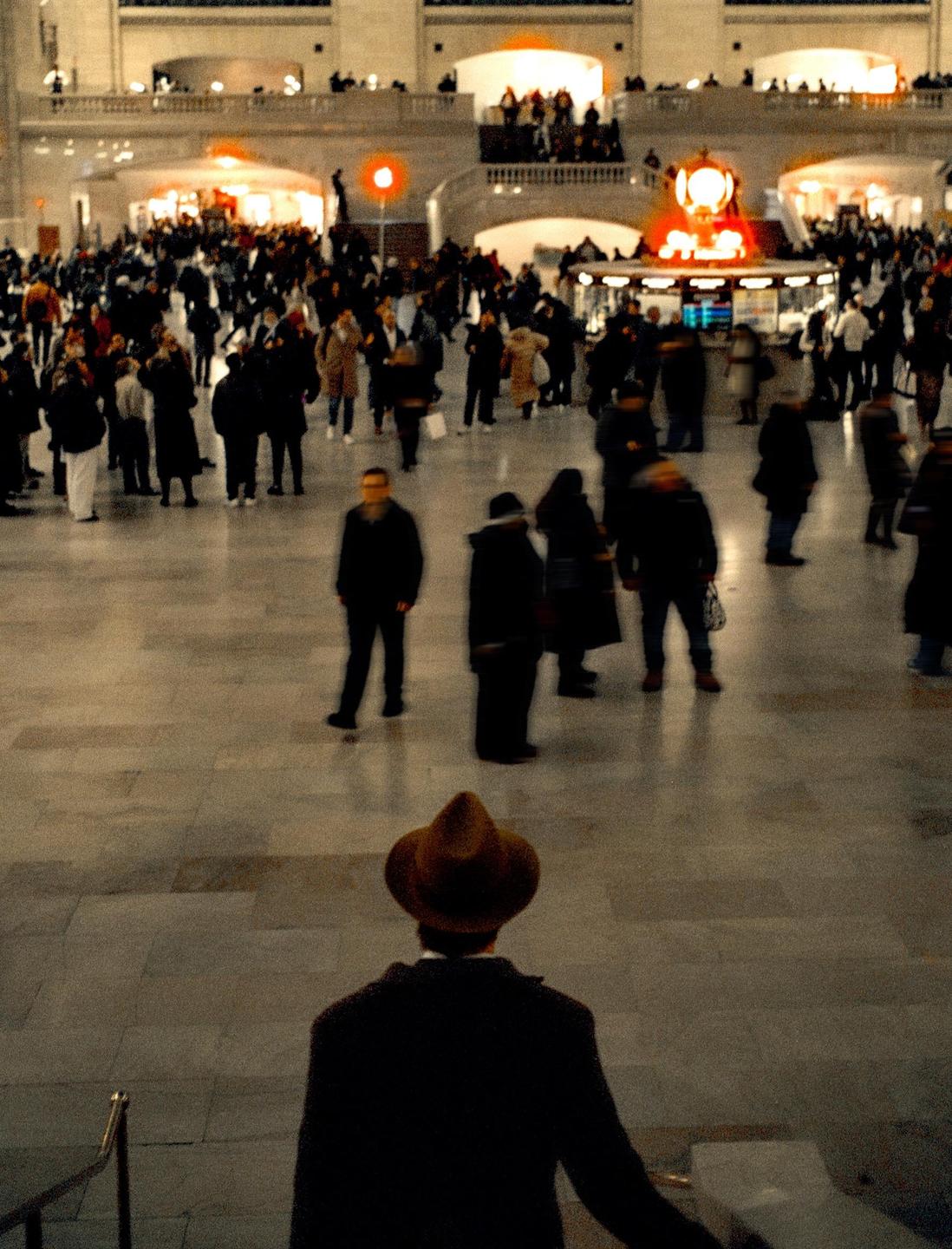 A man in a hat looks out over a busy train station concourse filled with people.