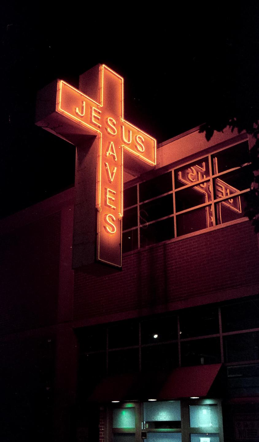 A neon sign in the shape of a cross with the words "Jesus Saves" is illuminated at night.