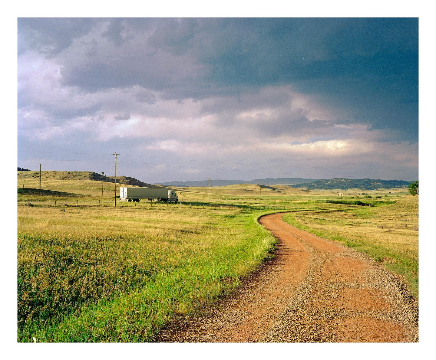 A dirt road curves through a grassy field under a cloudy sky, with a semi-truck parked in the distance.