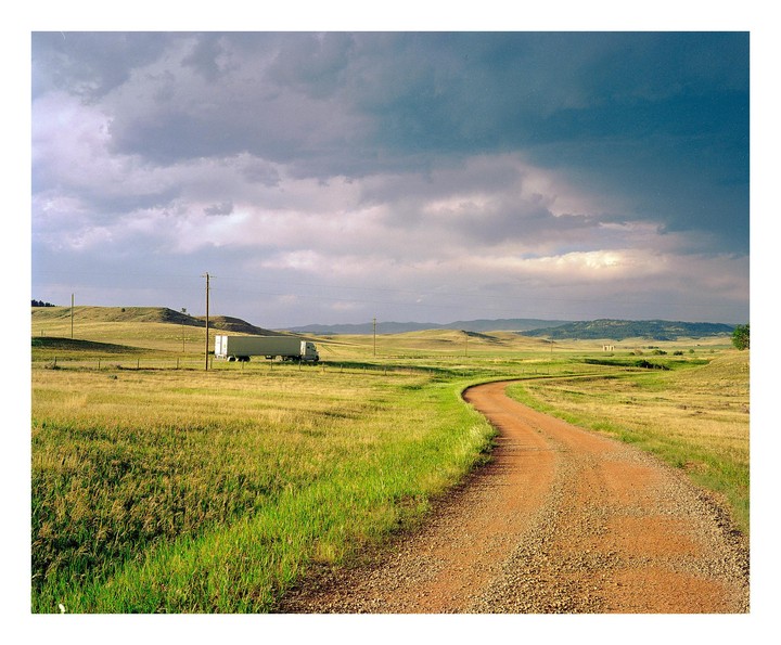 A dirt road curves through a grassy field under a cloudy sky, with a semi-truck parked in the distance.