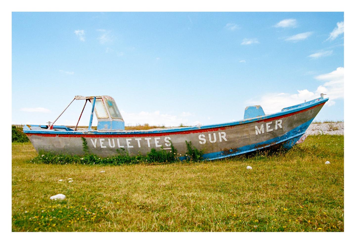An old boat with "VEULETTES SUR MER" written on its side sits in a grassy field under a blue sky.