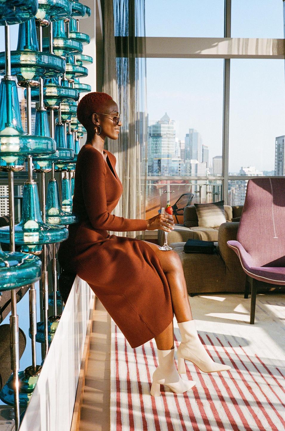 A woman in a brown dress sits on a ledge next to a large blue glass sculpture, holding a drink and looking out a window at a city skyline.