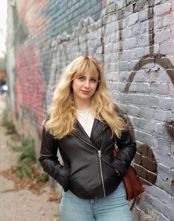 A woman with long blonde hair and a black leather jacket stands in front of a graffiti-covered brick wall.
