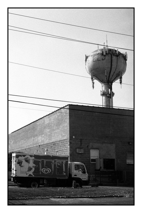 An ice cream truck is parked next to a brick building with a water tower and power lines above it.