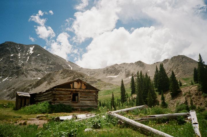 A wooden cabin sits in a grassy field with mountains and pine trees in the background under a blue sky with clouds.