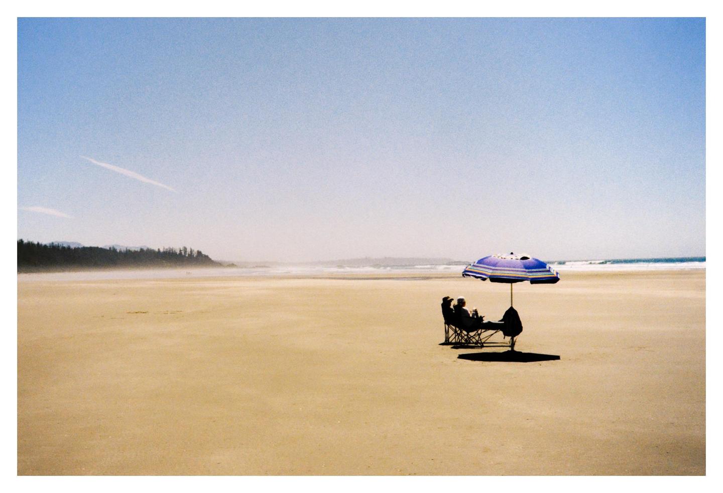 Two people sit under a purple and white striped umbrella on a vast sandy beach with a distant treeline and ocean waves.