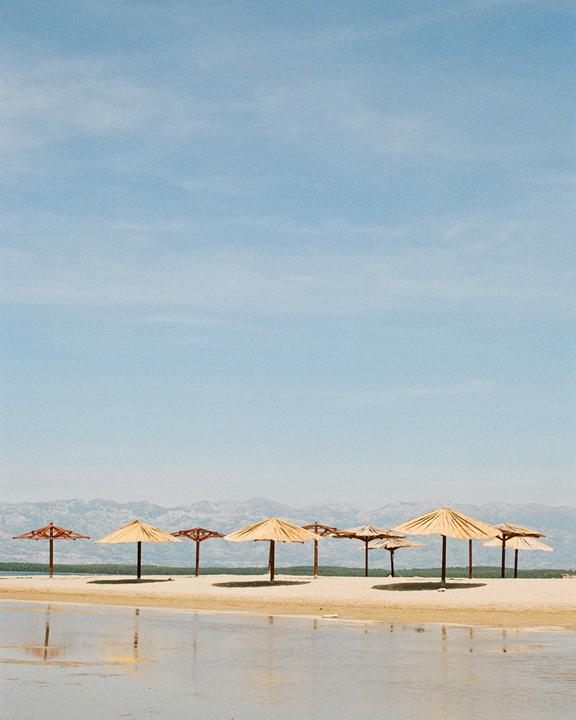 A row of straw beach umbrellas stands on a sandy beach with mountains in the background.