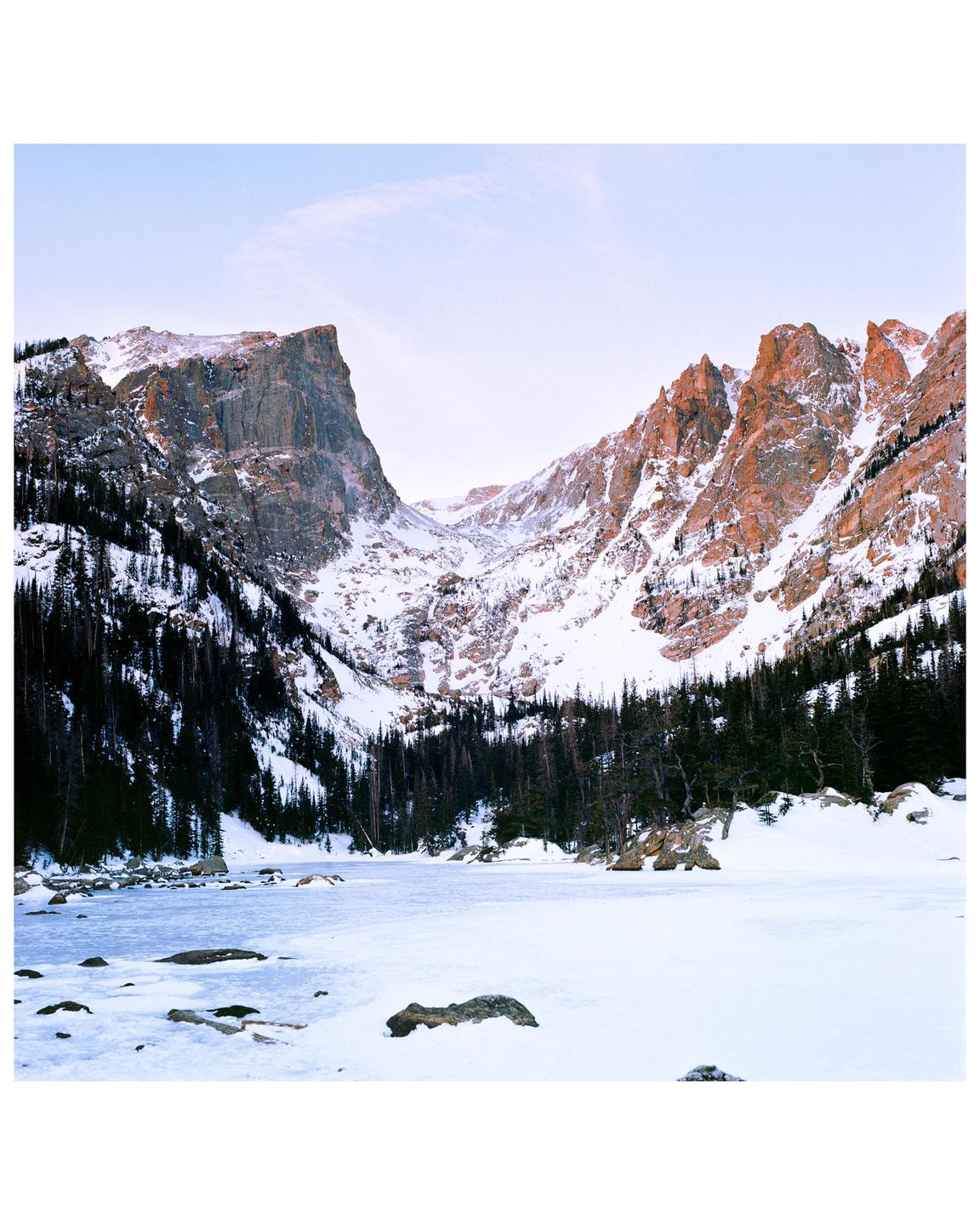 A frozen lake is surrounded by snow-covered mountains and pine trees under a pale sky.
