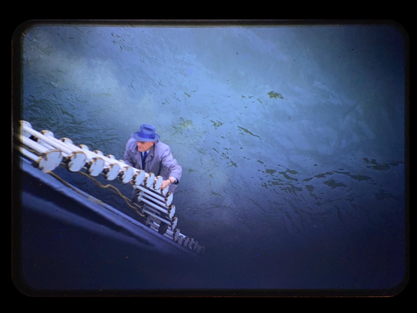 A man in a blue hat and coat climbs a ladder on the side of a boat, with water visible below him.