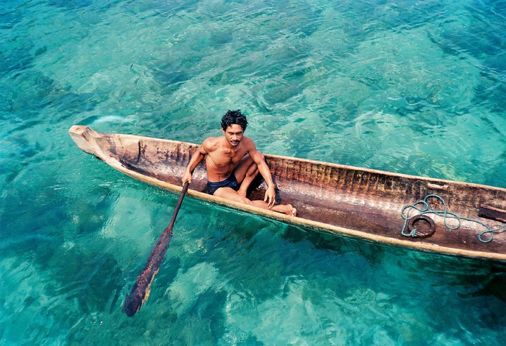 A man sits in a wooden boat with a paddle in clear turquoise water.