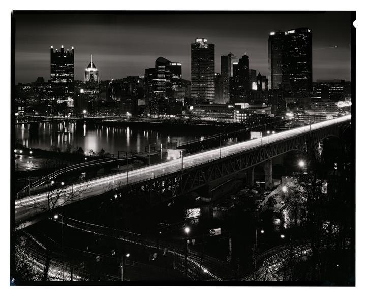 A city skyline with tall buildings and a bridge over a river is illuminated at night.