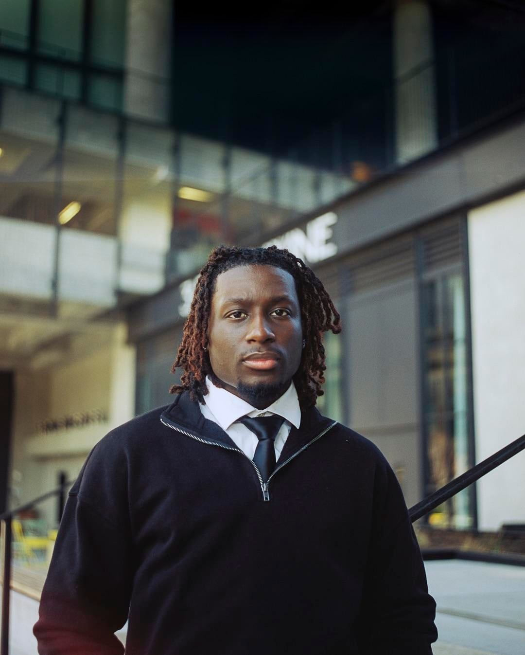 A man with dreadlocks, a white shirt, and a black tie and sweater stands in front of a modern building.