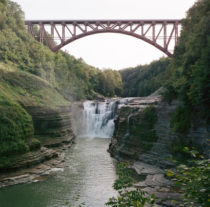A waterfall cascades into a river, framed by a bridge overhead and lush green cliffs.