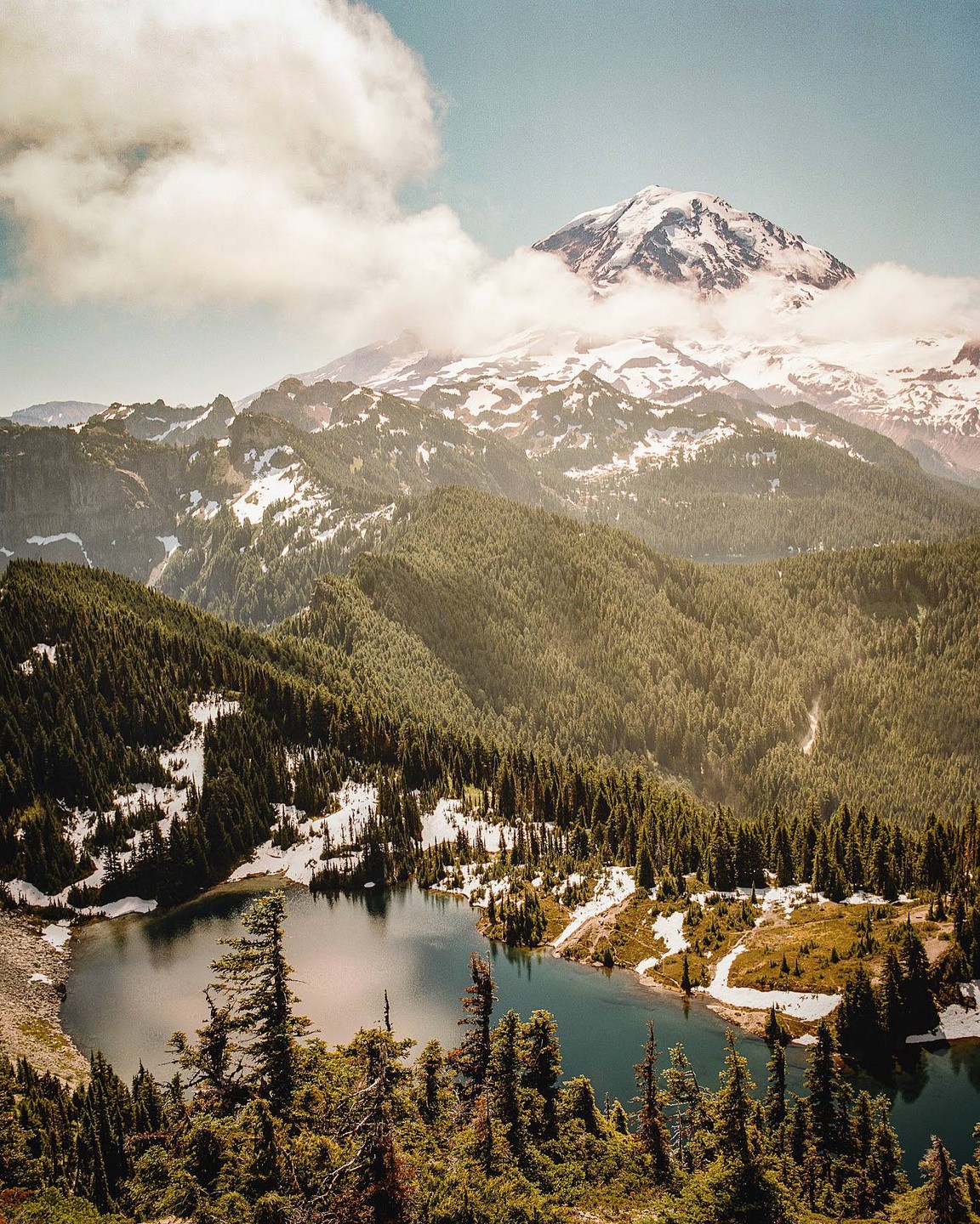 A snow-capped mountain peak rises above a forest and a lake.