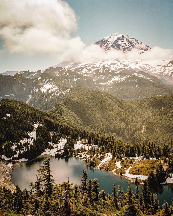 A snow-capped mountain peak rises above a forest and a lake.