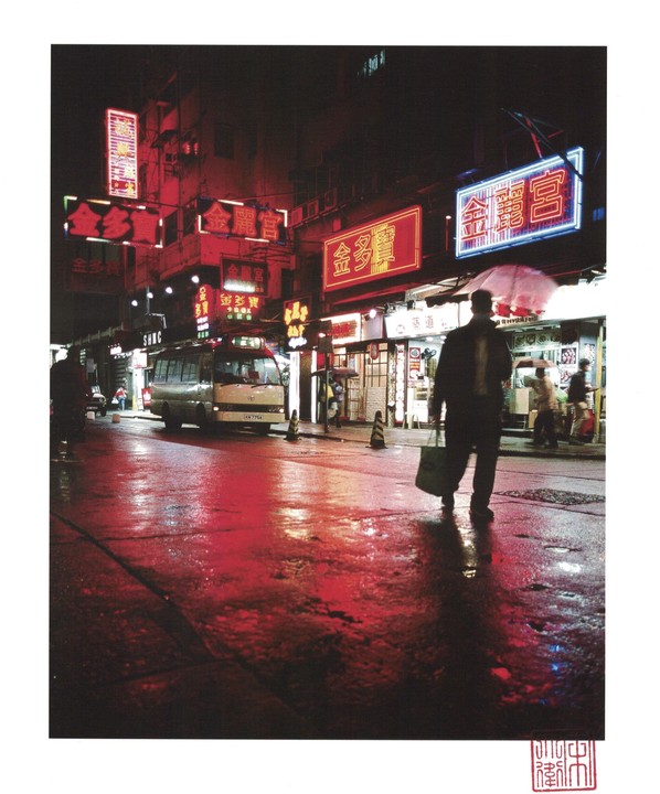 A man walks down a wet street at night, illuminated by red and blue neon signs.