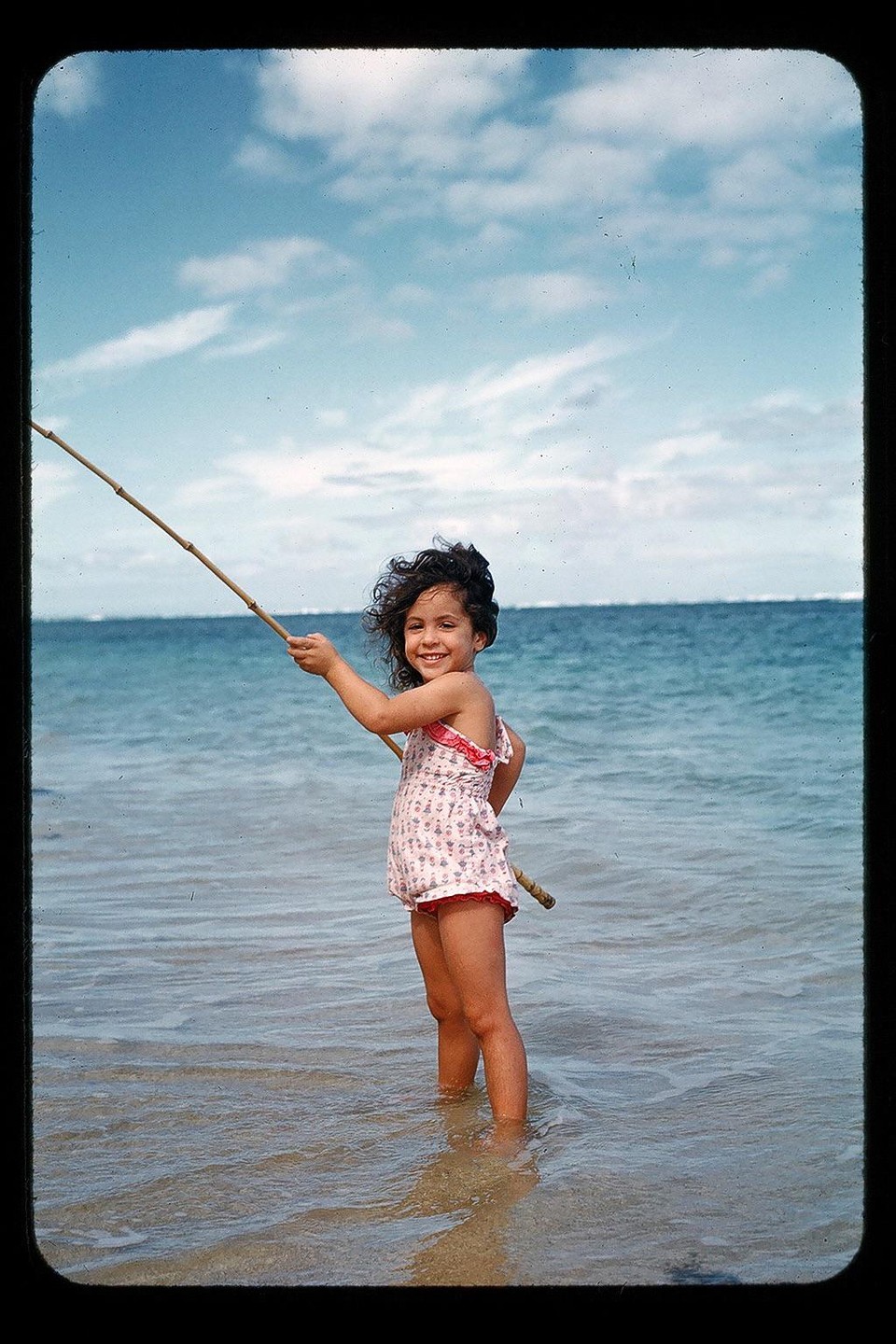 A young girl in a swimsuit stands in the ocean holding a fishing pole.