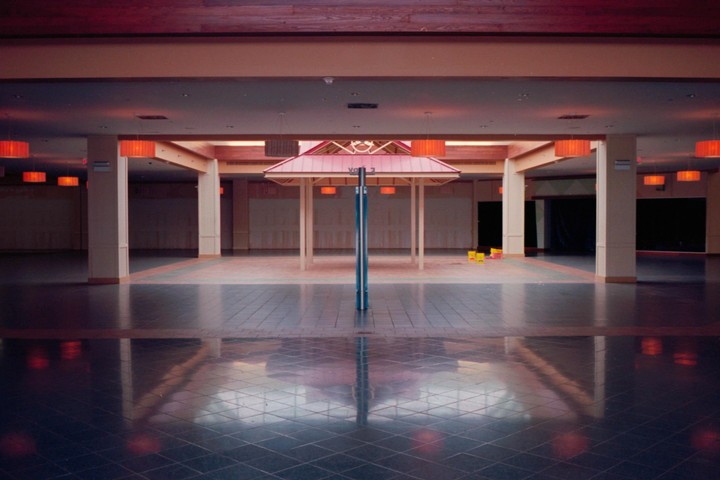An empty mall interior features a central kiosk with a red roof and numerous orange pendant lights reflecting on the tiled floor.