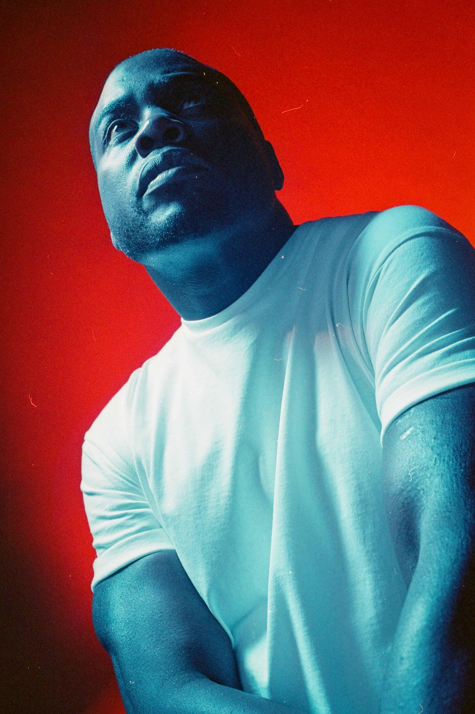 A man in a white t-shirt looks up against a red background, with his skin appearing blue.