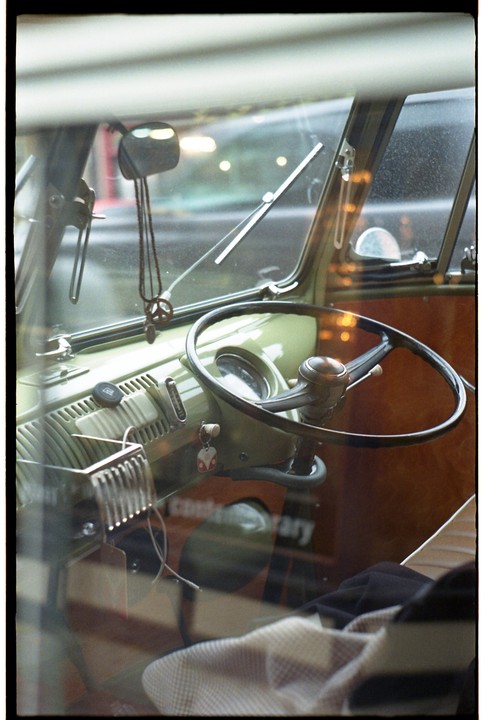 The interior of a vintage green van is visible through its windshield, showing the steering wheel, dashboard, and a peace sign hanging from the rearview mirror.