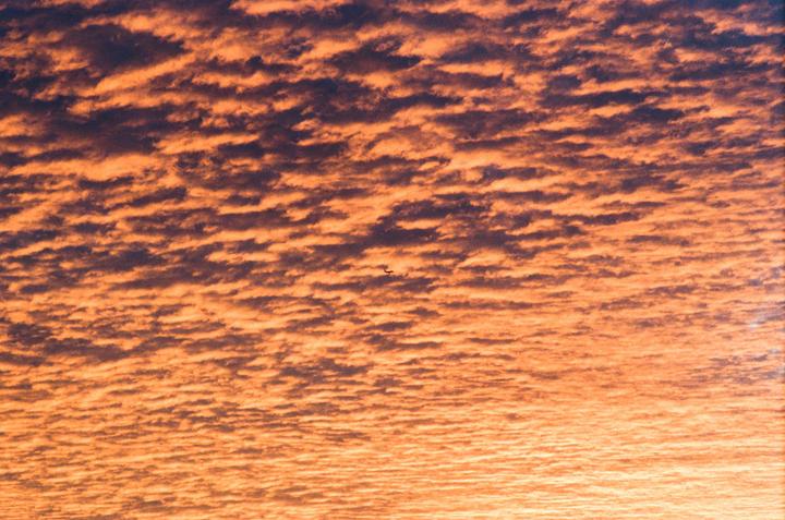 A small airplane flies through a sky filled with orange and dark clouds during sunset.