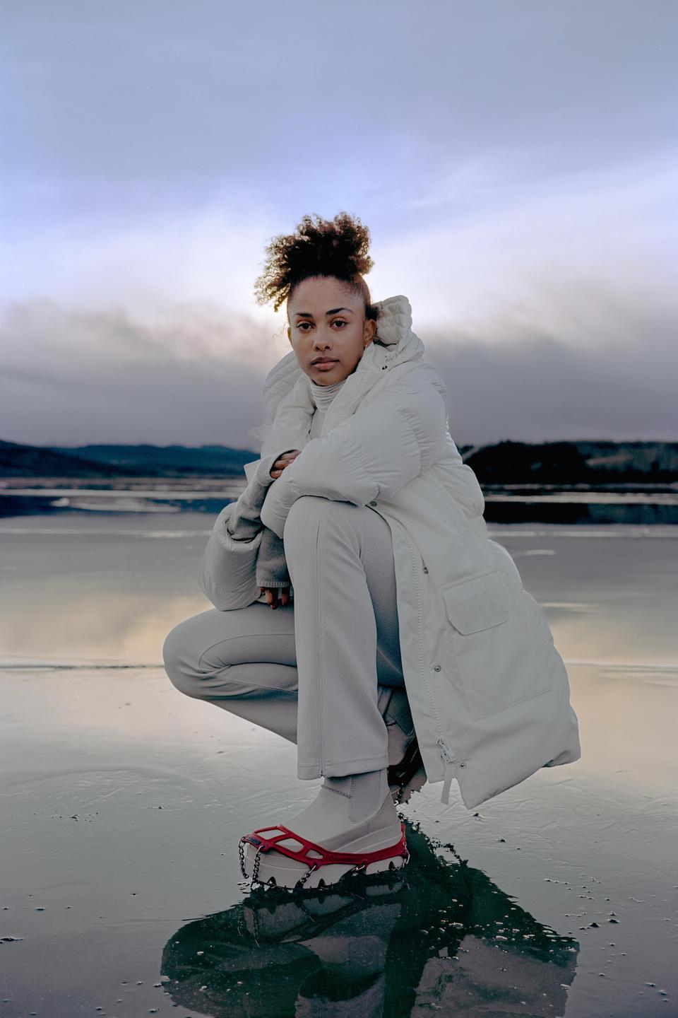 A woman wearing a white coat and red ice cleats crouches on a wet surface with a body of water and mountains in the background.